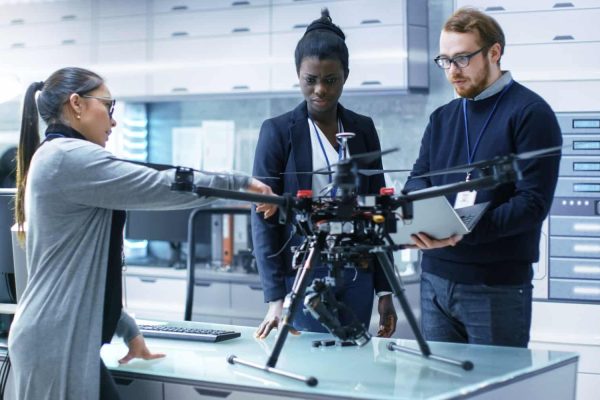 Multi Ethnic Team of Young Female and Male Engineers Working on Modern Drone Prototype. They're in a Bright High-Tech Laboratory.